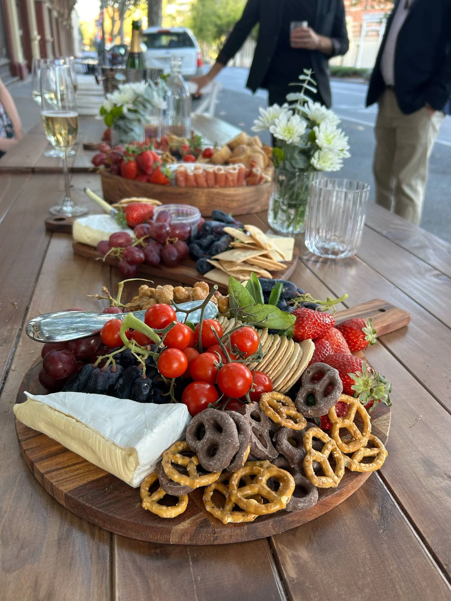 A cheese and fruit platter with grapes, strawberries, cherry tomatoes, pretzels, crackers, and cheese on a wooden serving board. In the background, there are wine glasses, a flower arrangement, and people standing around outdoor dining table.