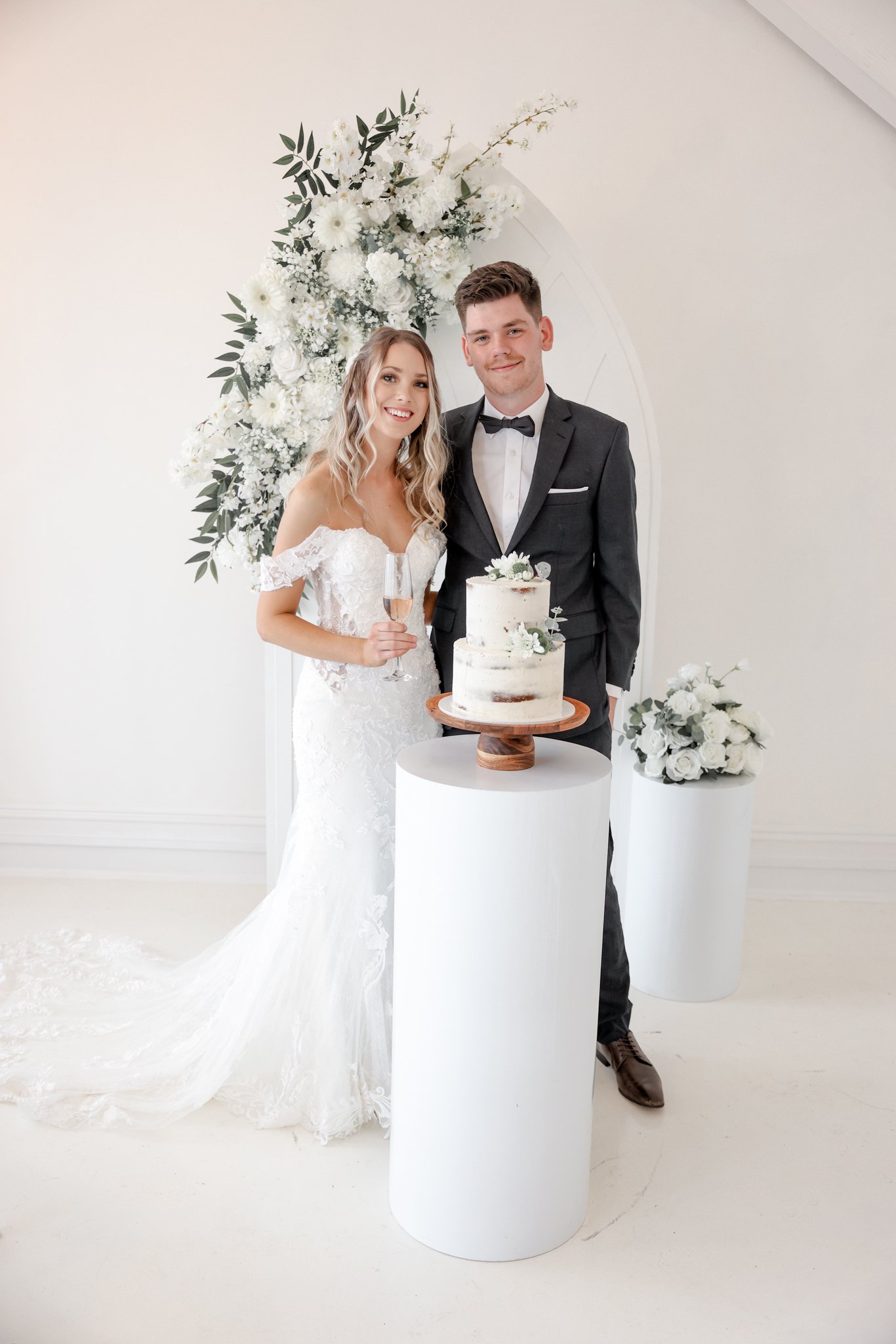 A bride and groom at their wedding celebration standing behind a wedding cake on a white pedestal with floral arrangements in the background.