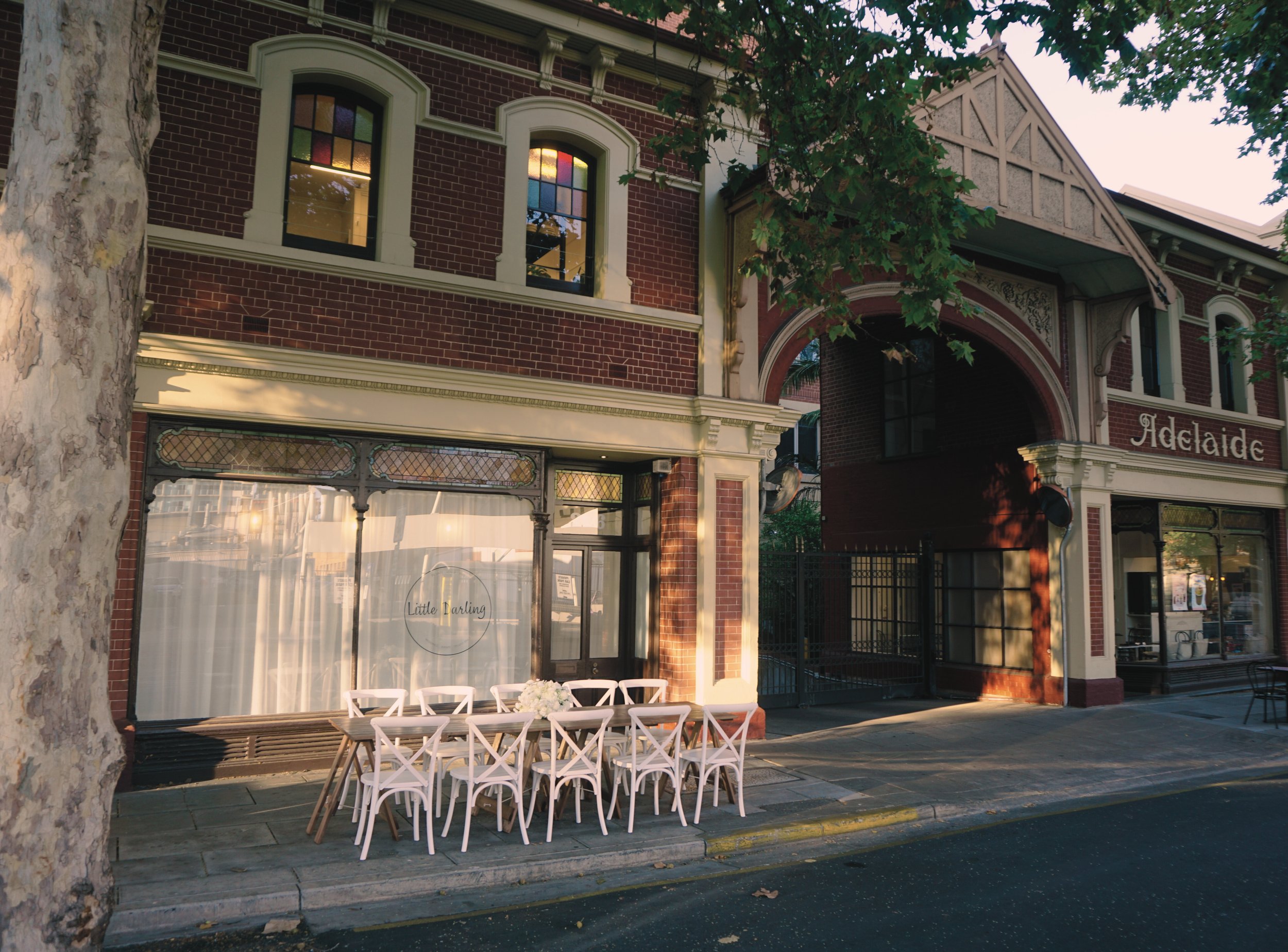 A street view of a brick building with large arched windows and a sign reading "Adelaide." In front of the building, there is a table set for outdoor dining with nine white chairs and a flower arrangement, illuminated by warm sunlight. A tree partial