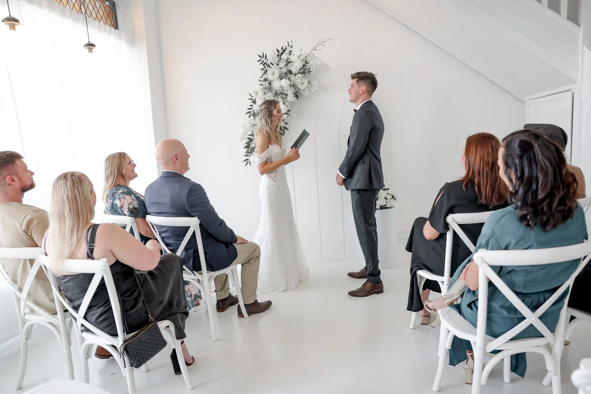 A bride and groom stand facing each other during a wedding ceremony in a bright room with white walls and a floral arrangement behind them. The bride wears a white lace wedding dress, and the groom wears a gray suit. Guests are seated on white chairs