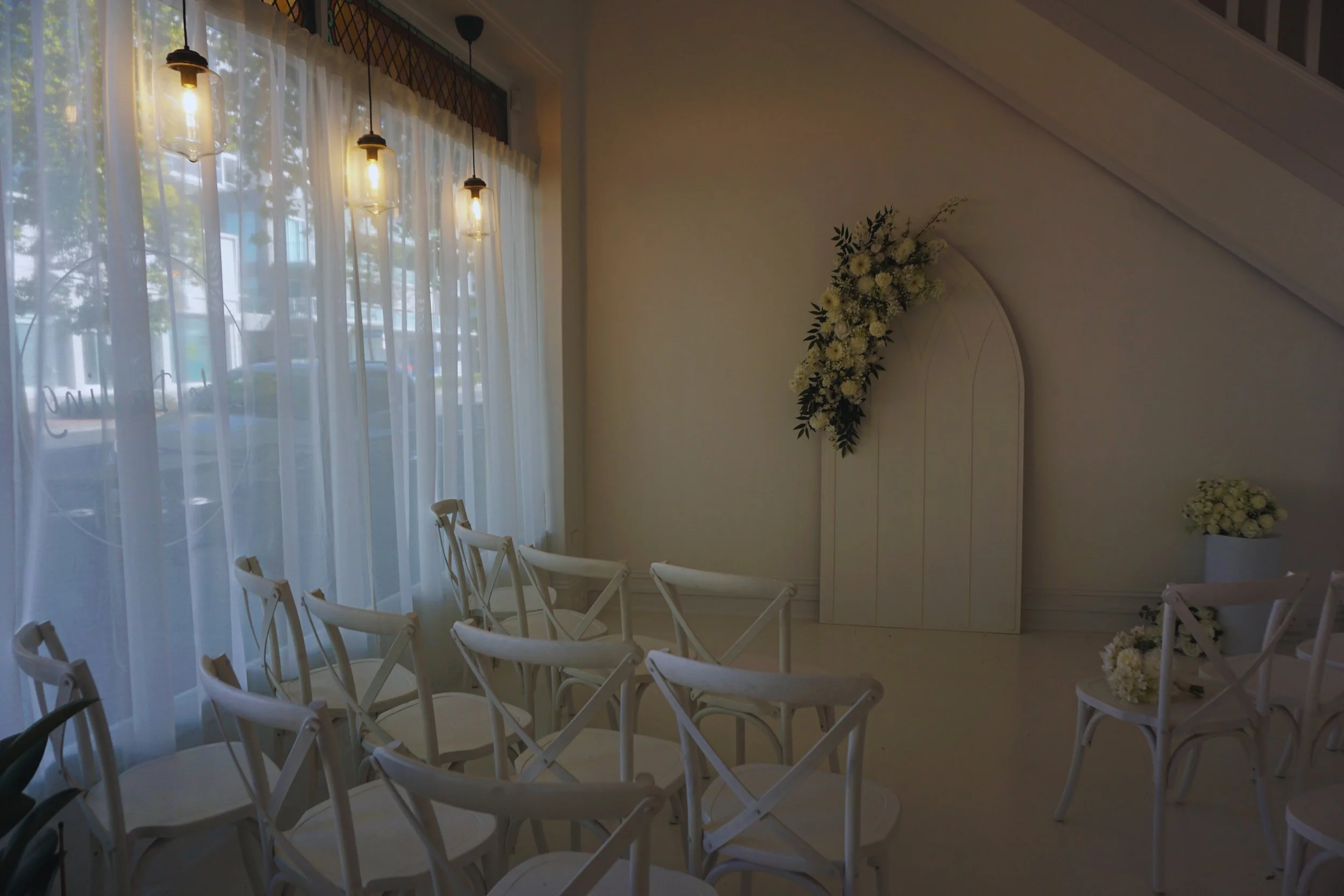 Interior of a wedding or event space with white chairs, floral arrangements, and large windows with white curtains, decorated for a ceremony.
