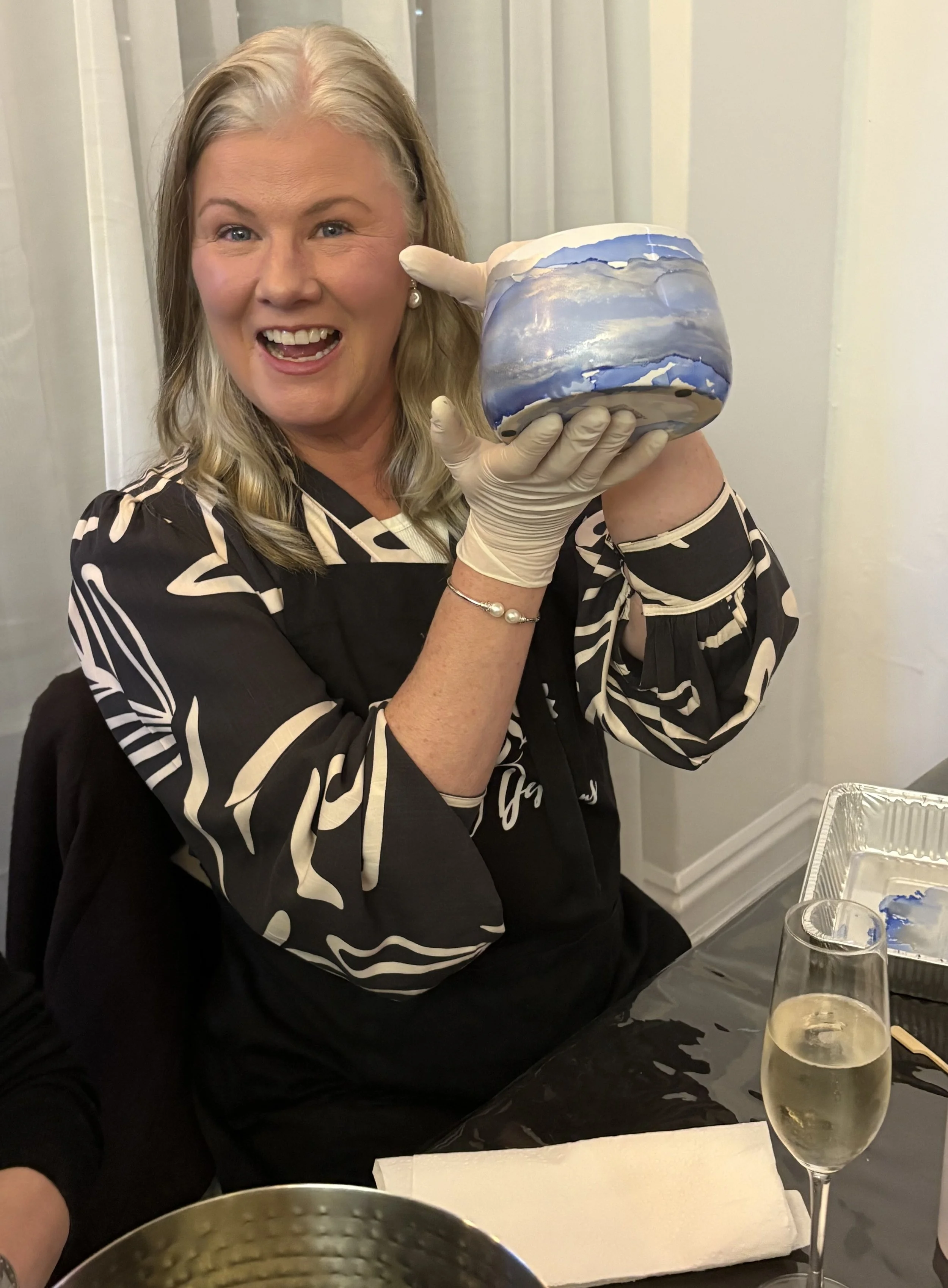 A woman with blonde hair, wearing a black and white patterned blouse, is smiling and holding a marbled blue and white cake. She is wearing gloves and pearl earrings, and sitting at a table with a glass of champagne nearby.
