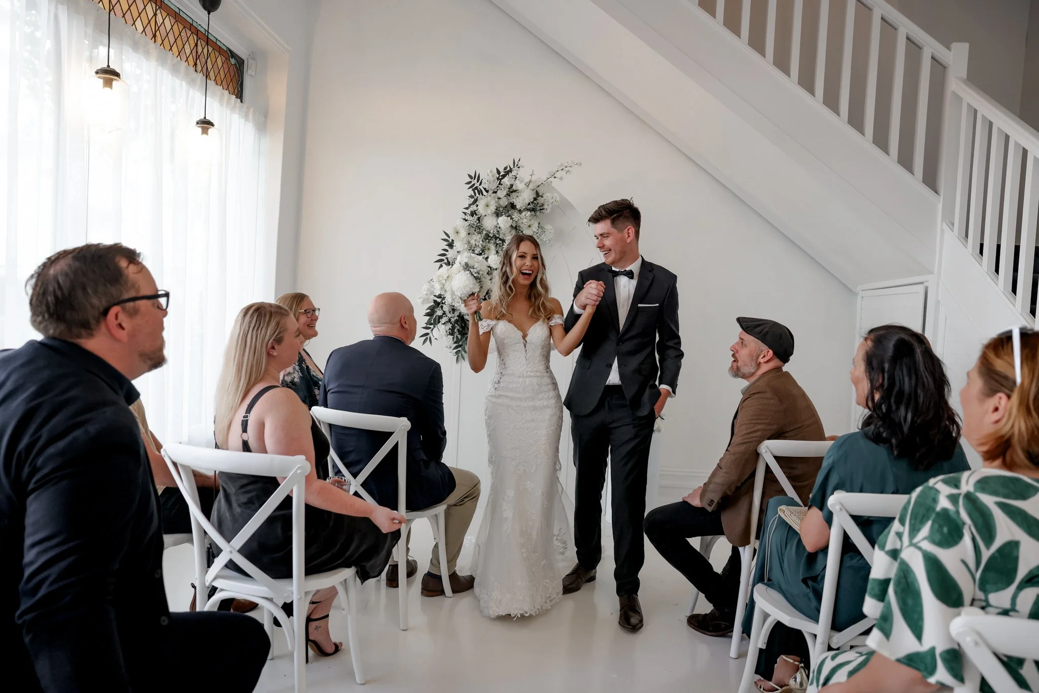 A bride and groom are standing together at their wedding ceremony, holding hands and smiling, surrounded by seated guests.