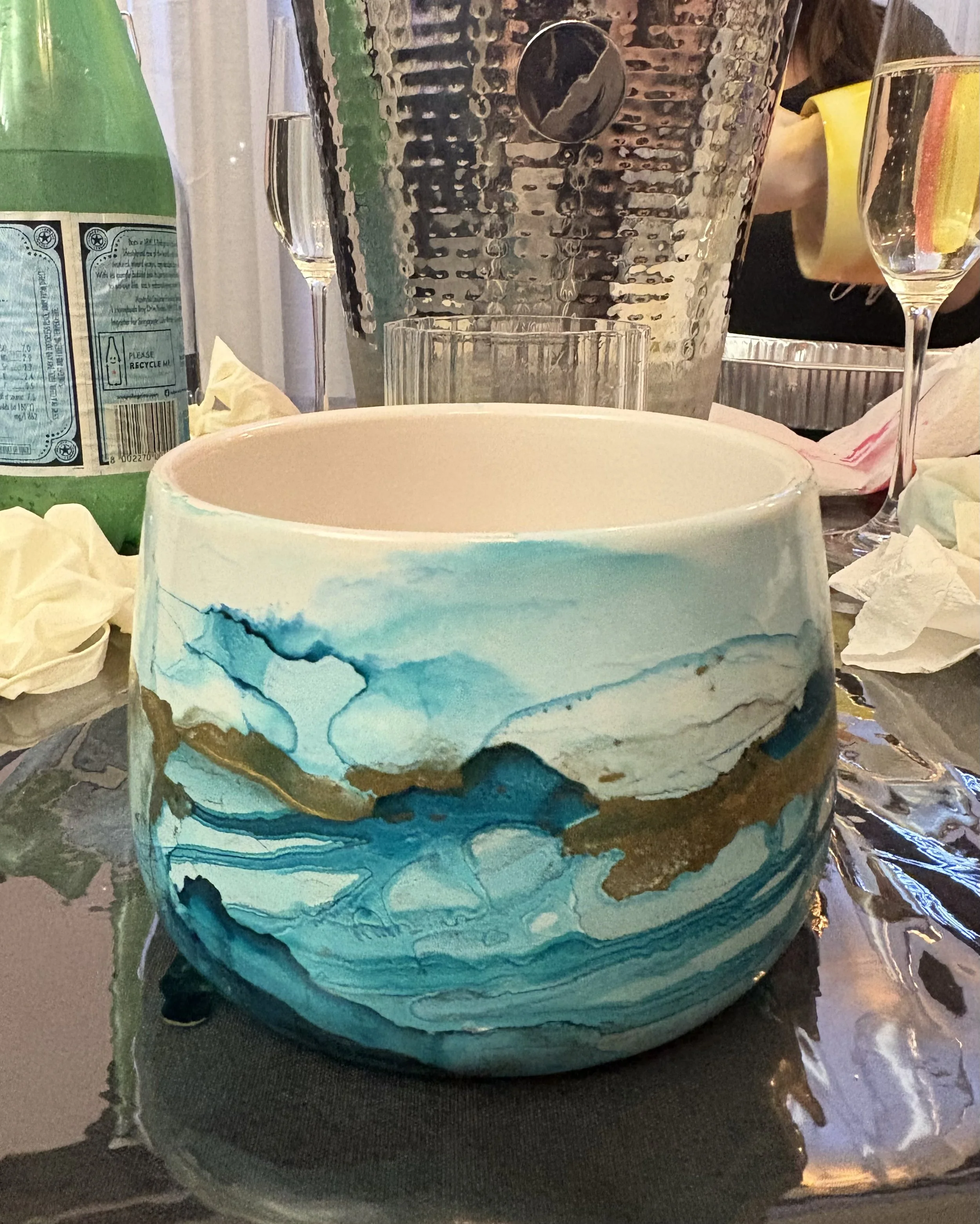 Round decorative bowl with blue, white, and gold abstract design on a table surrounded by crumpled paper and drink glasses.