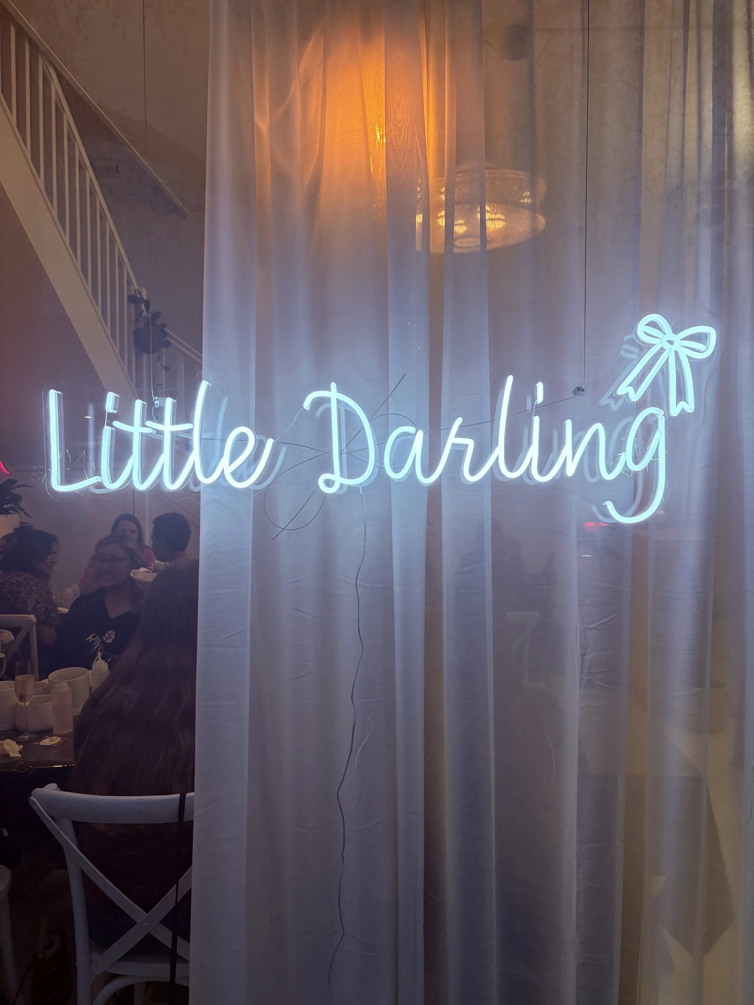 Neon sign reading 'Little Darlin' with a bow graphic on a curtain, behind a party scene with people dining.