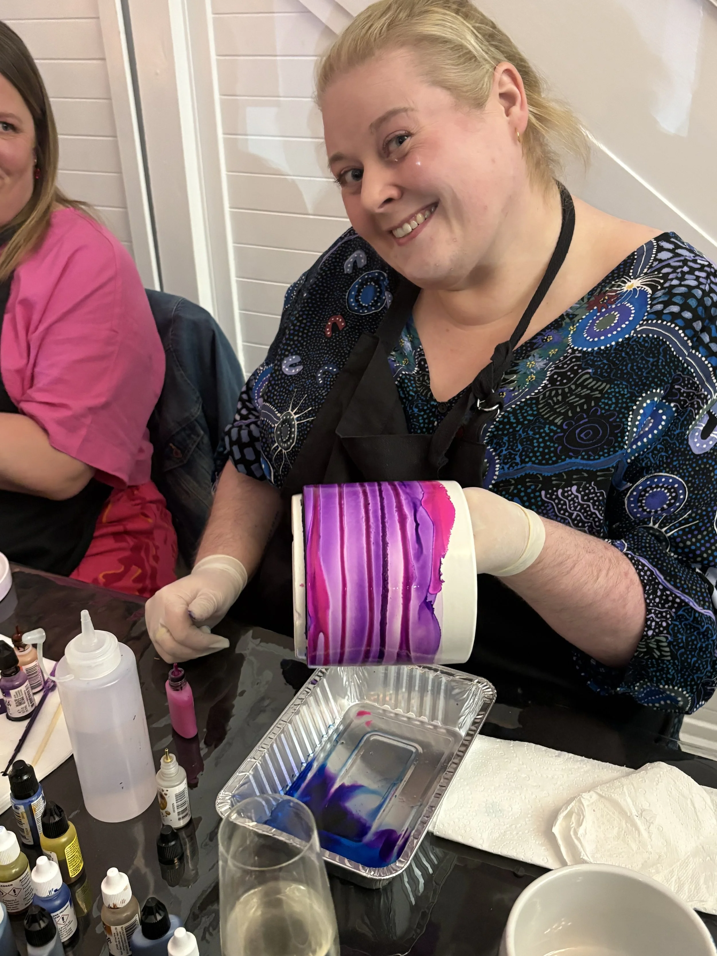 Woman in a black apron and latex gloves holding a white mug with purple and pink dye flowing down its sides, surrounded by bottles of dye, a plastic squeeze bottle, paper towels, and a glass of clear liquid, with another person partially visible in t