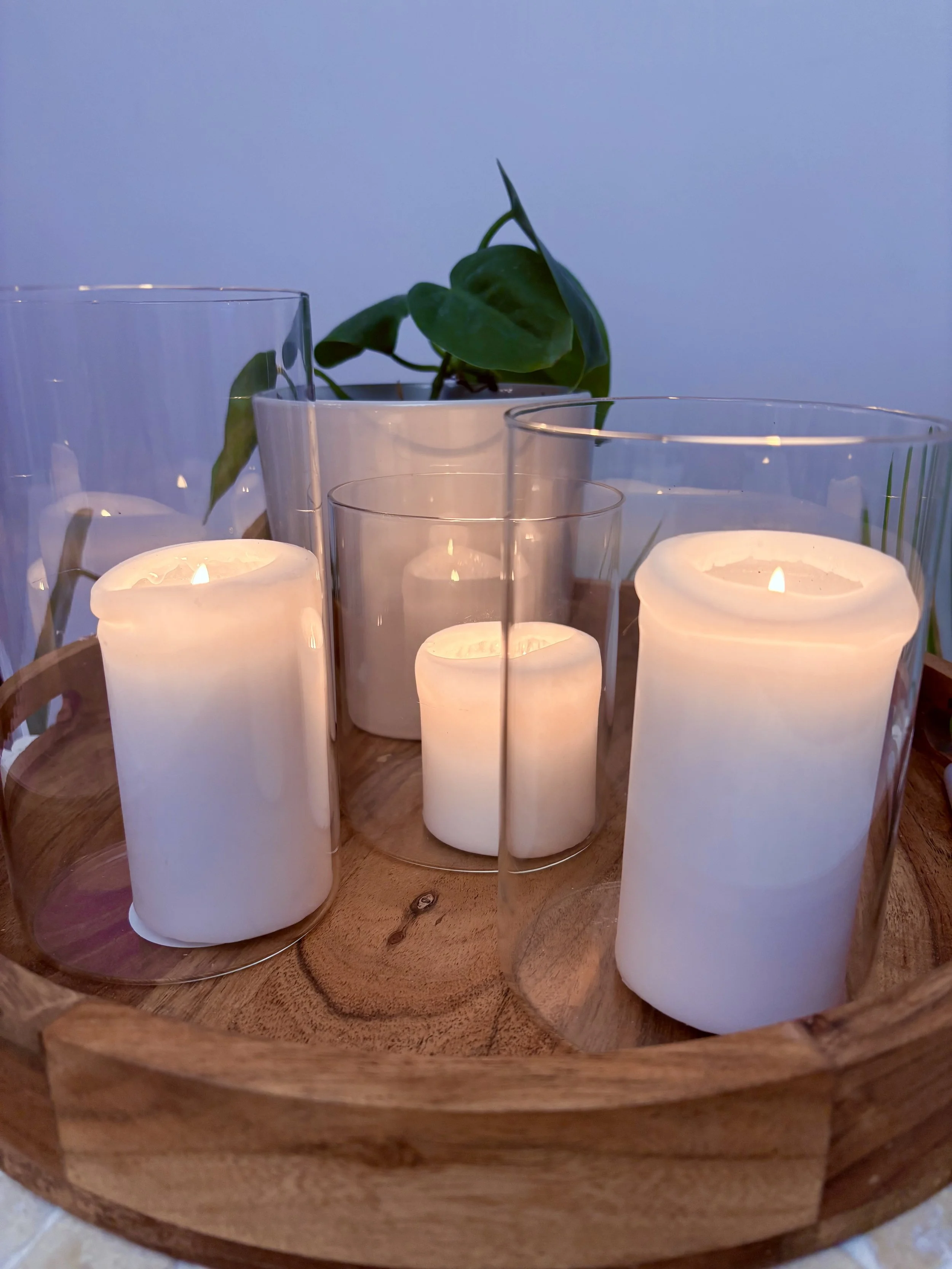 Four white candles in glass holders on a wooden tray with a green potted plant in the background.