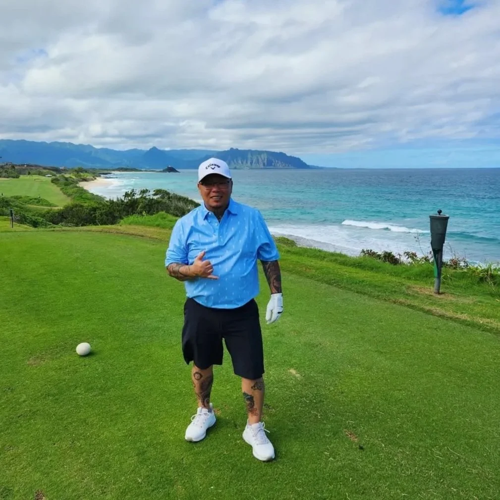 Man standing on a golf course near the coastline, wearing a blue shirt, shorts, white golf glove, and cap, with mountains and ocean in the background.