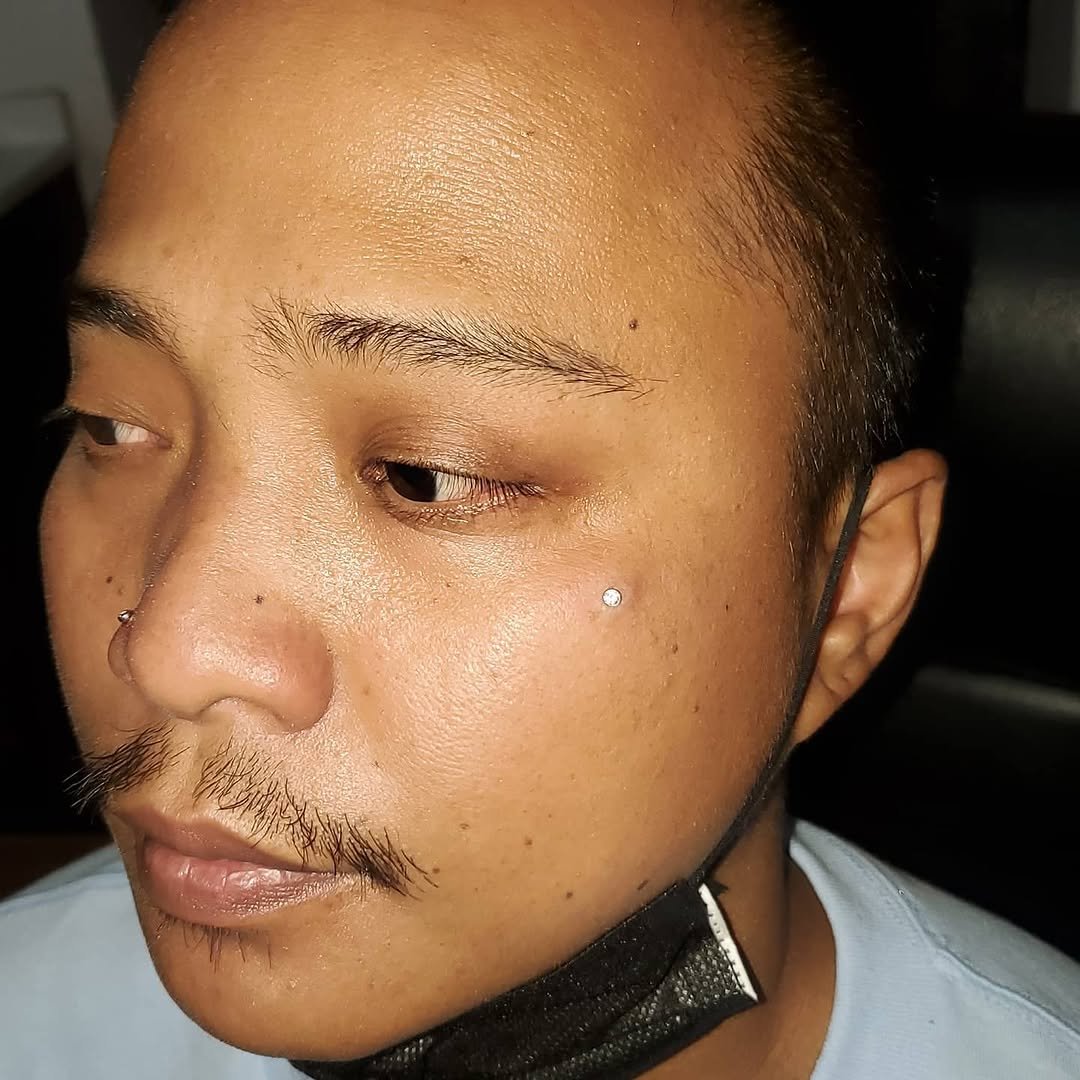 Close-up of a person's face with a focus on the left side, showing smooth skin, minimal makeup, a nose piercing, a small diamond earring, and a partially pulled-down black face mask.