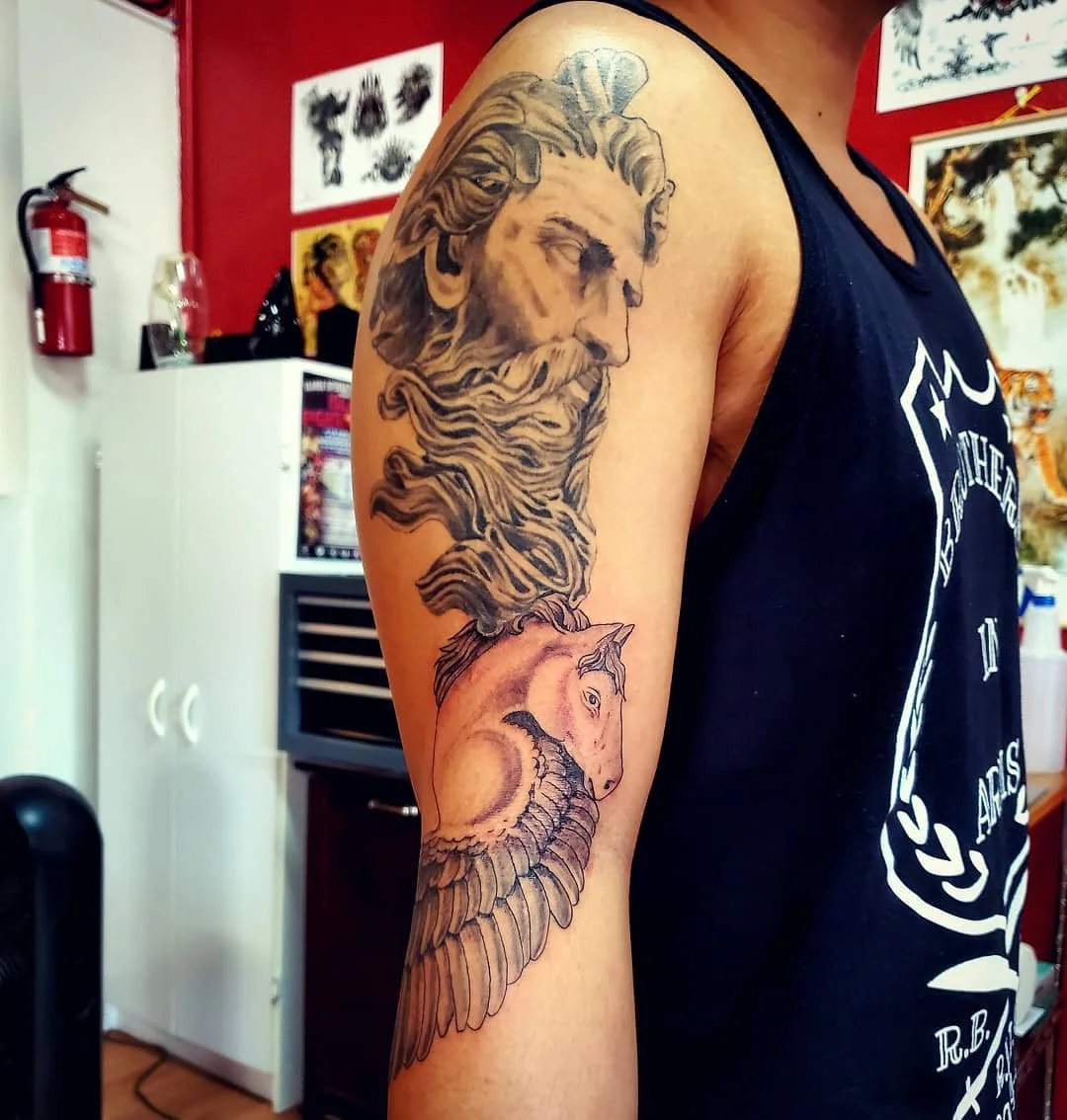 Tattoo of a bearded man's face with flowing hair and a unicorn's head on an upper arm.