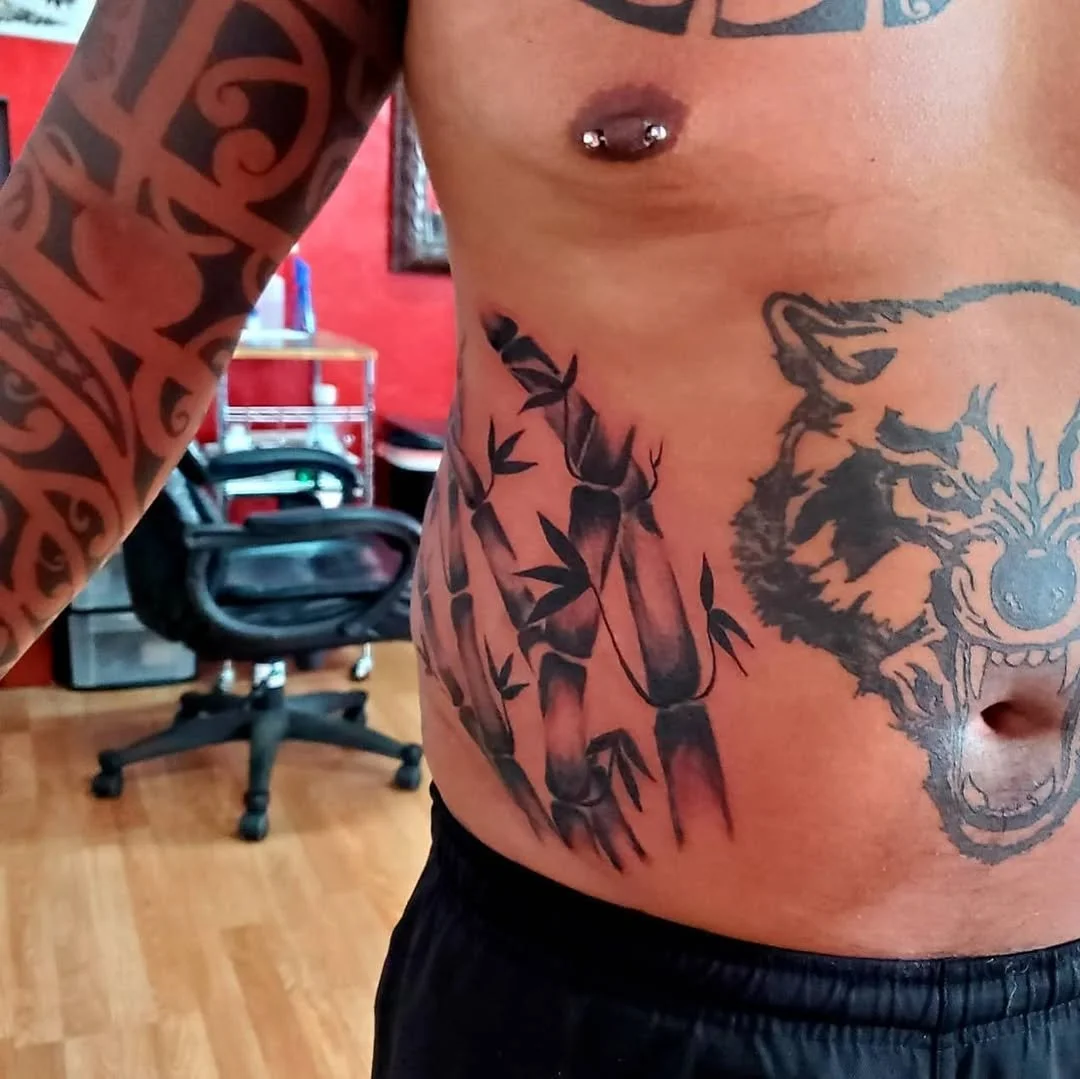 Close-up of a tattooed male torso with tribal and animal designs, including a wolf or bear face, and a nipple piercing, in a room with a wooden floor, black chair, and red walls.