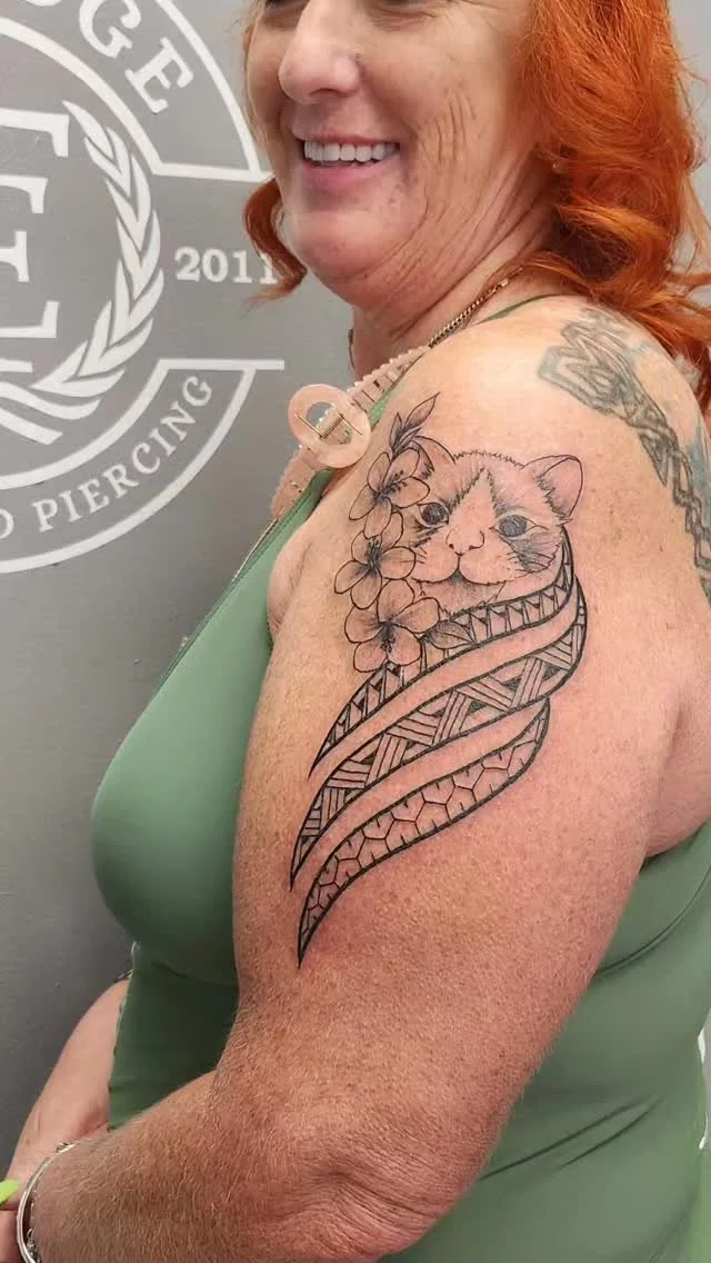 A woman with red hair smiling, showing a tattoo of a cat's face surrounded by flowers and tribal patterns on her upper arm.