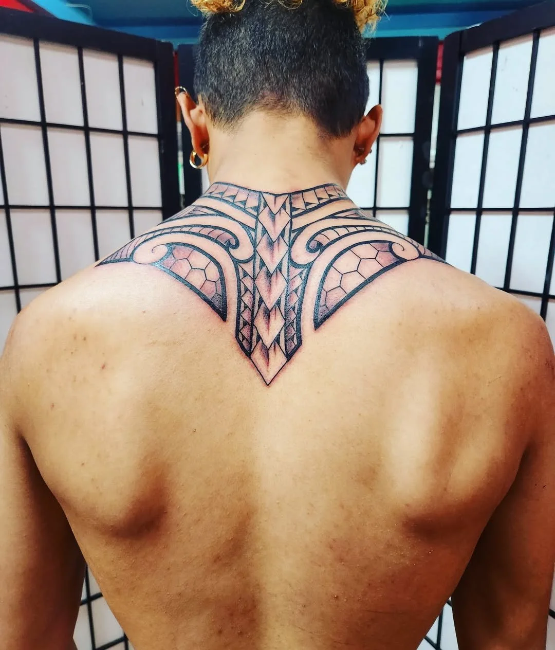 Back view of a person with a detailed black and gray tribal-style tattoo on their upper back and shoulders, in front of a black and white folding privacy screen.