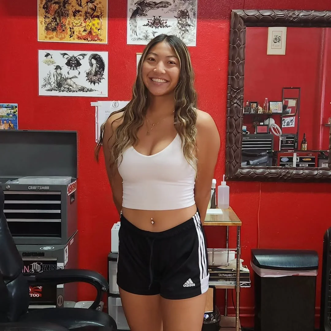 A young woman with long wavy hair and a belly button piercing, smiling while standing in a tattoo studio with red walls, framed artwork, and a large mirror.