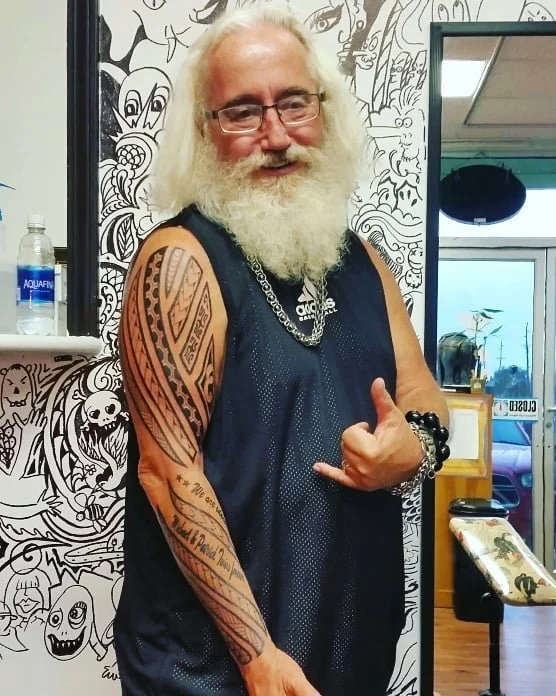A man with long white hair and a beard, wearing glasses, a black sleeveless athletic shirt, and jewelry, posing indoors with a thumb gesture in front of a black-and-white mural.