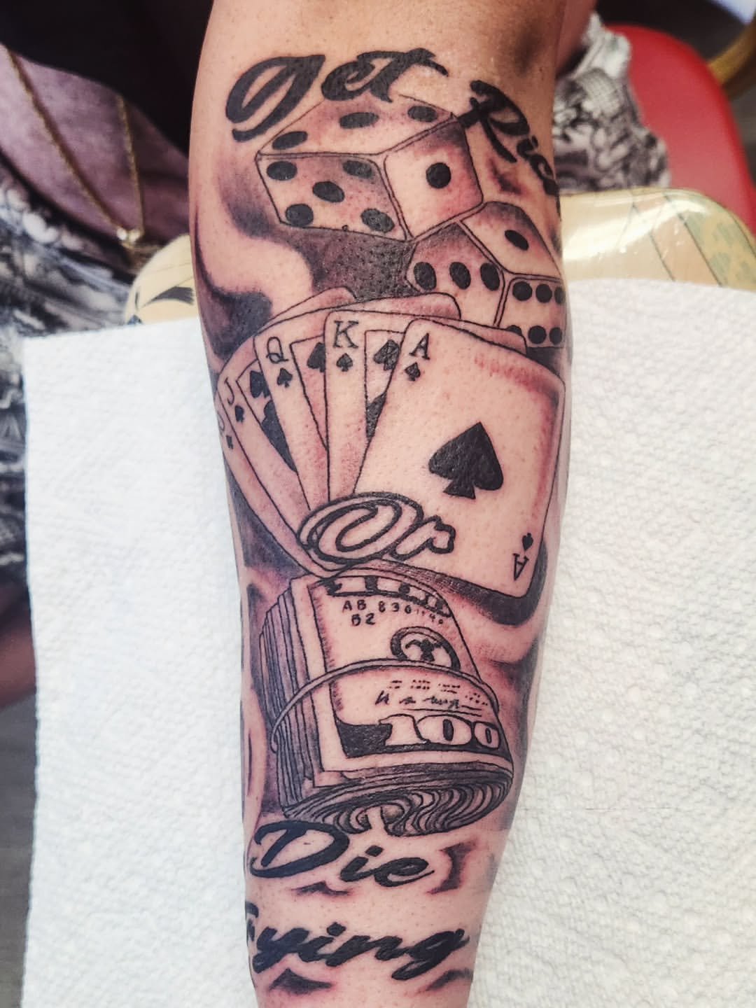 Tattoo of gambling elements, including dice, playing cards (Ace, King, Queen, Jack of spades), and a rolled-up bill with the words 'Die' and 'Dying' written on it.