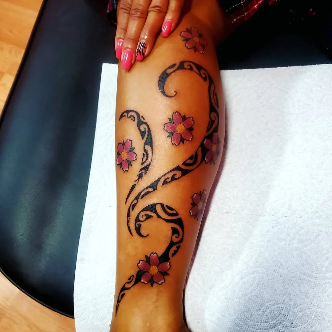 Freshly done tattoo of black swirling tribal patterns with pink cherry blossom flowers on a person's leg.