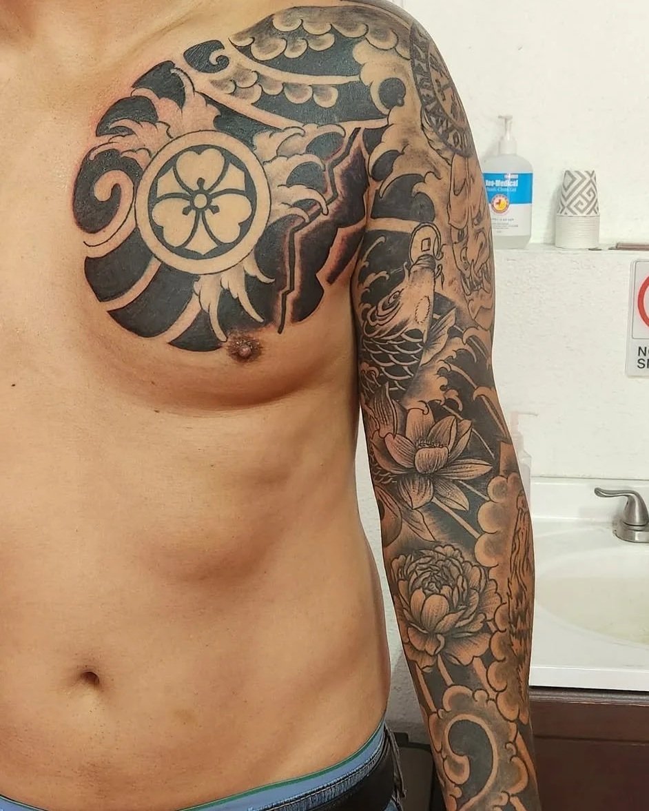 Partial view of a person's upper torso, showing extensive black and gray traditional tattoo sleeve with floral and wave motifs on the right arm and chest, with a background of a bathroom with soap dispenser and paper cups.