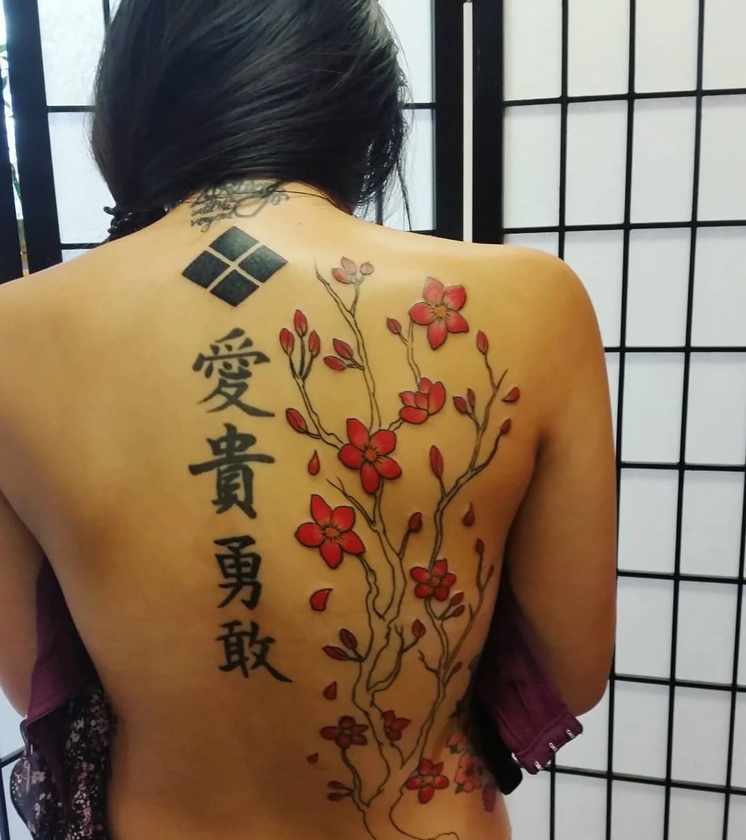 A woman with a large tattoo of cherry blossoms and Chinese characters on her back, standing in front of a black and white folding screen.