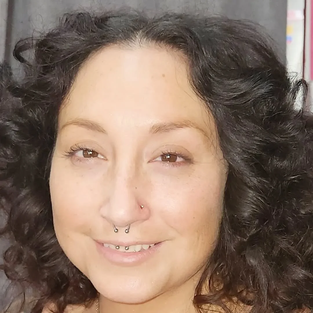 Close-up of a woman with curly dark hair, light skin, and multiple facial piercings, smiling at the camera.