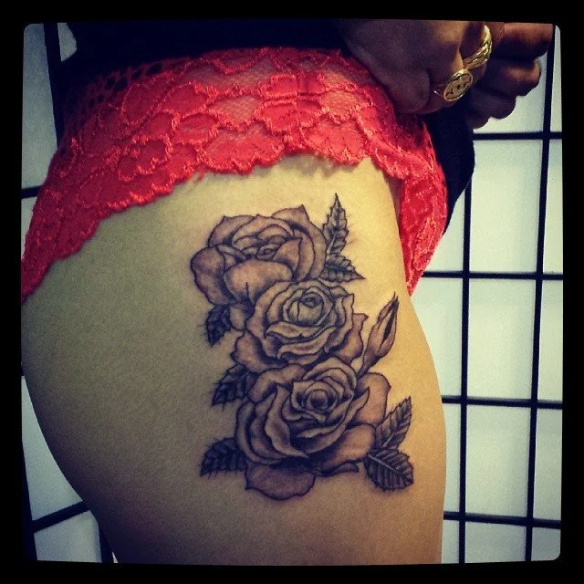 Tattoo of three roses with leaves on the upper thigh, with red lace underwear and hand with rings visible in the background.