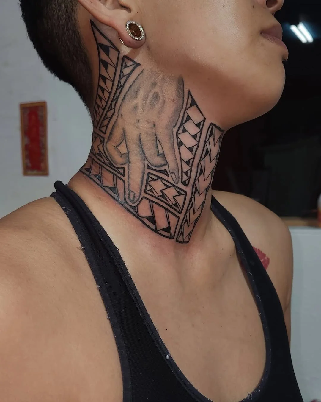 Close-up of a person's neck and lower face showing a black geometric tattoo with intricate lines and patterns, and a hand tattoo with extended fingers. The person has earrings and is wearing a black tank top.