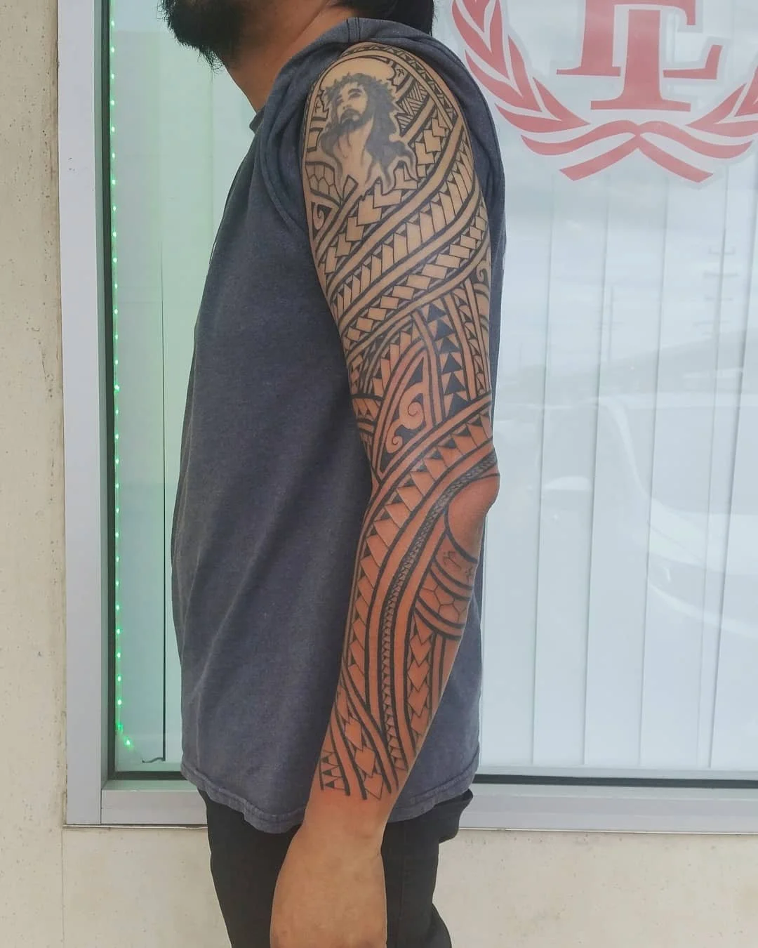 A man with a Polynesian-style tribal tattoo sleeve on his right arm, wearing a sleeveless hoodie, standing indoors in front of a glass wall with a logo.