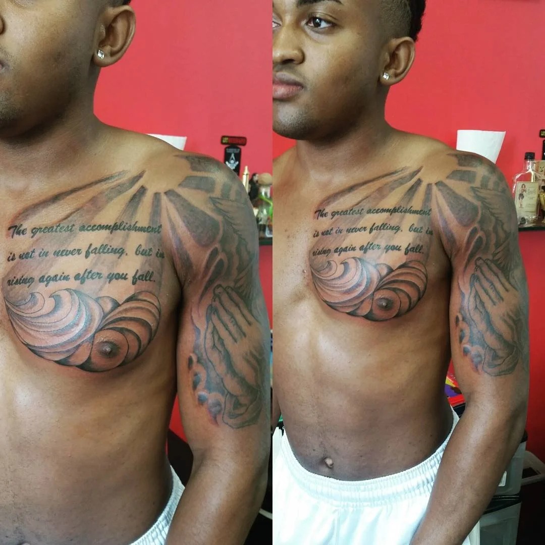 A shirtless man with a shoulder tattoo of a quote and artistic design, standing in front of a red wall with various items on a shelf behind him.