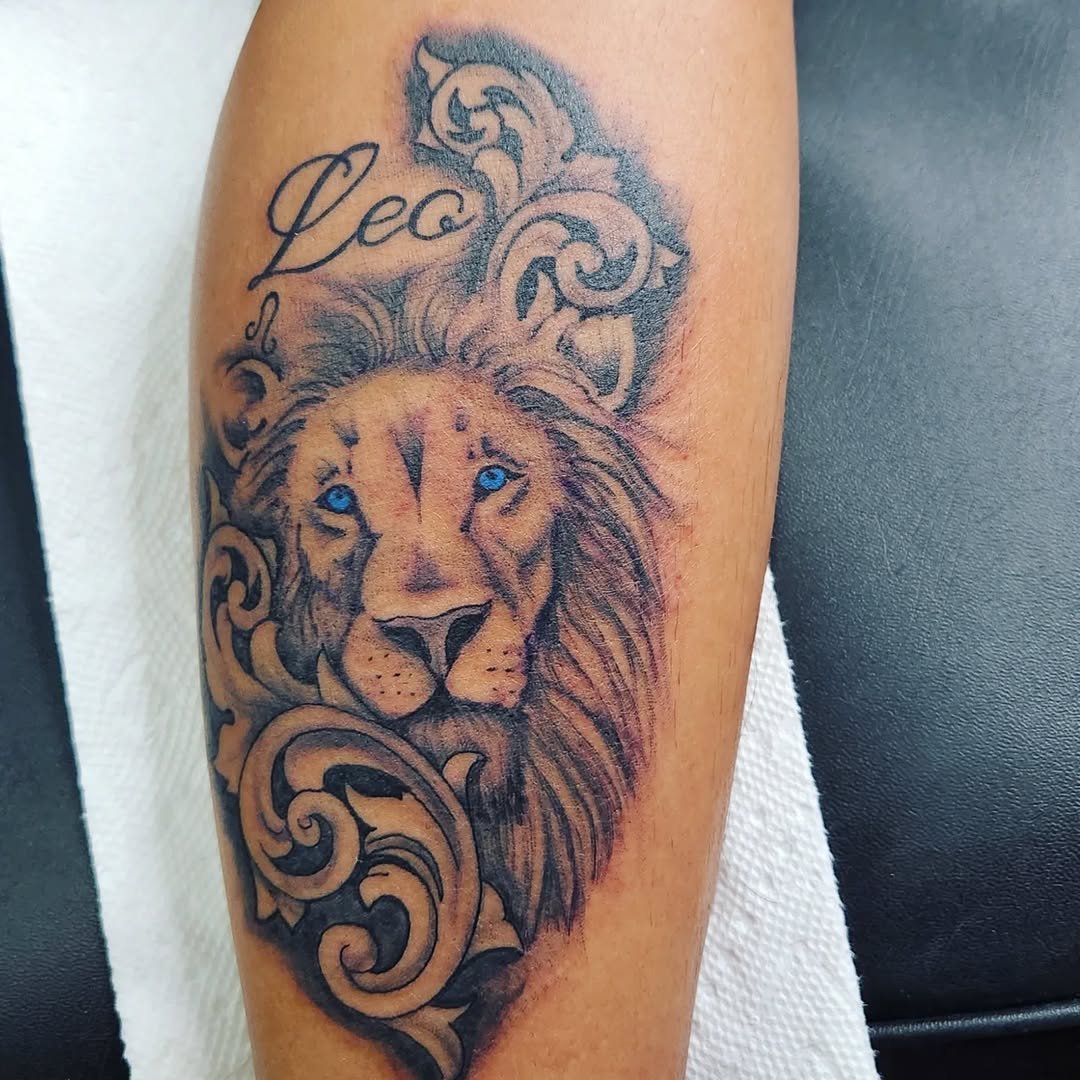 Tattoo of a lion's face with blue eyes, surrounded by decorative scrollwork and feathers, with the word 'Leo' written above.