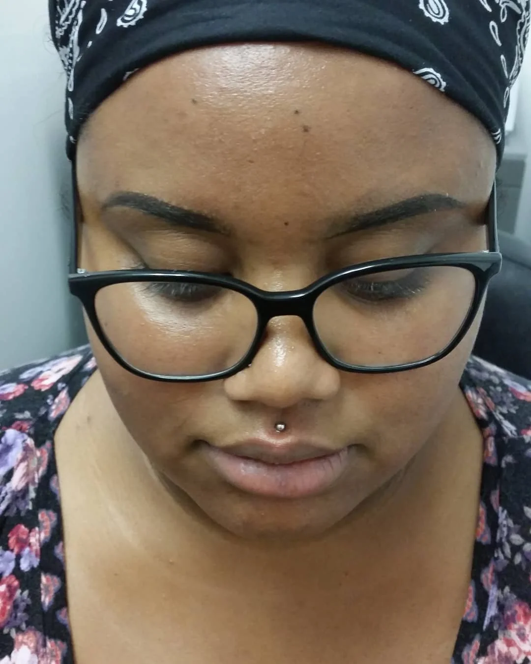 Close-up of woman wearing black glasses and headscarf, with a piercing above her upper lip, looking downward.