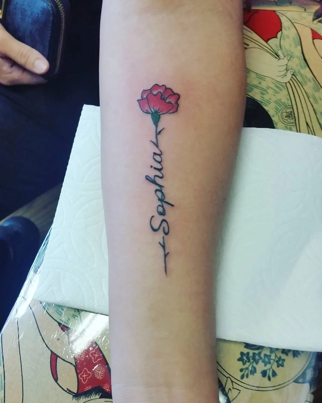 Tattoo of a red carnation flower with green stem and leaves and the word 'Sophie' written vertically in cursive on the person's forearm.