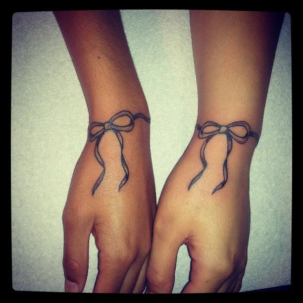 Two hands held side by side, each with a tattoo of a simple black outline of a bow on the wrist.