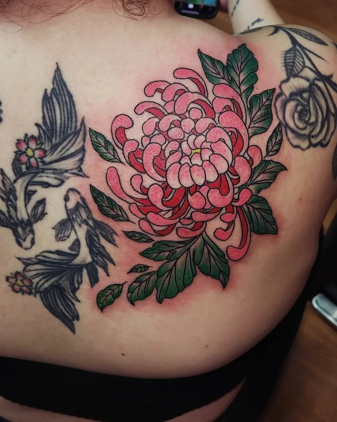 Colorful pink and red flower tattoo on person's shoulder, with green leaves and black-and-white floral tattoos nearby.