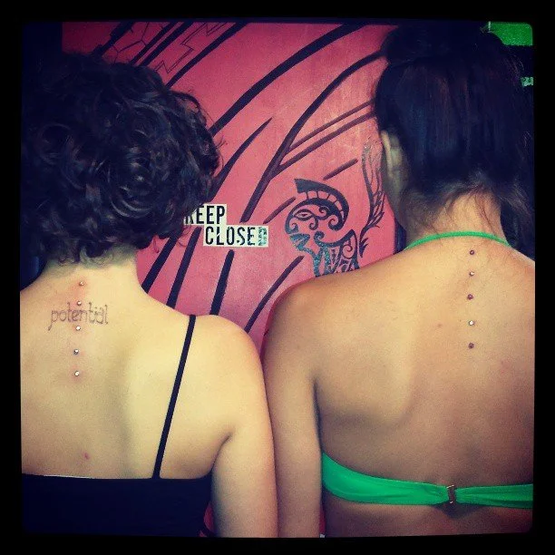 Two women with back piercings facing a colorful wall with a graffiti design, one with a tattoo that says 'potential' on her back.