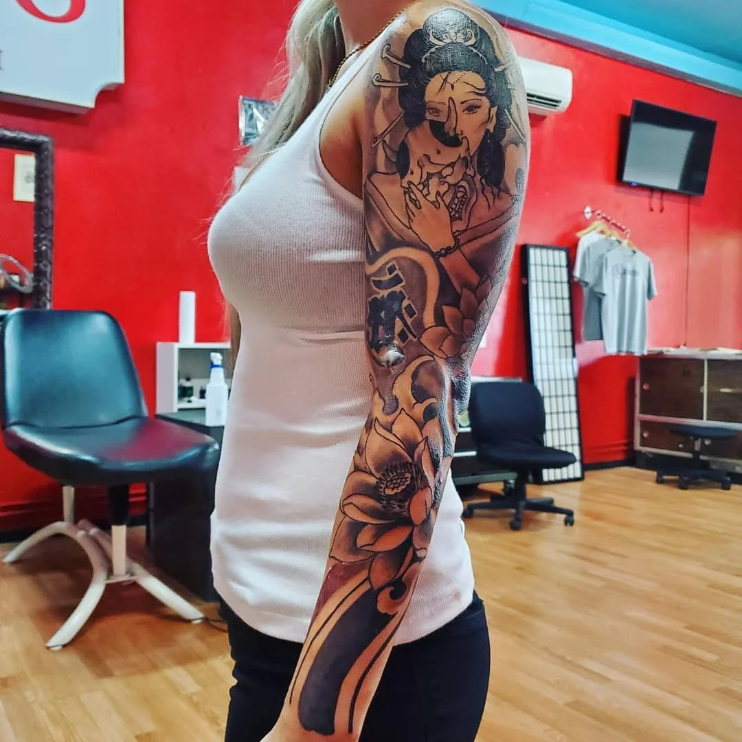A woman with a detailed black and gray tattoo sleeve on her left arm featuring a woman in traditional Asian attire, flowers, and abstract designs, standing in a tattoo studio with a red wall and various furniture.