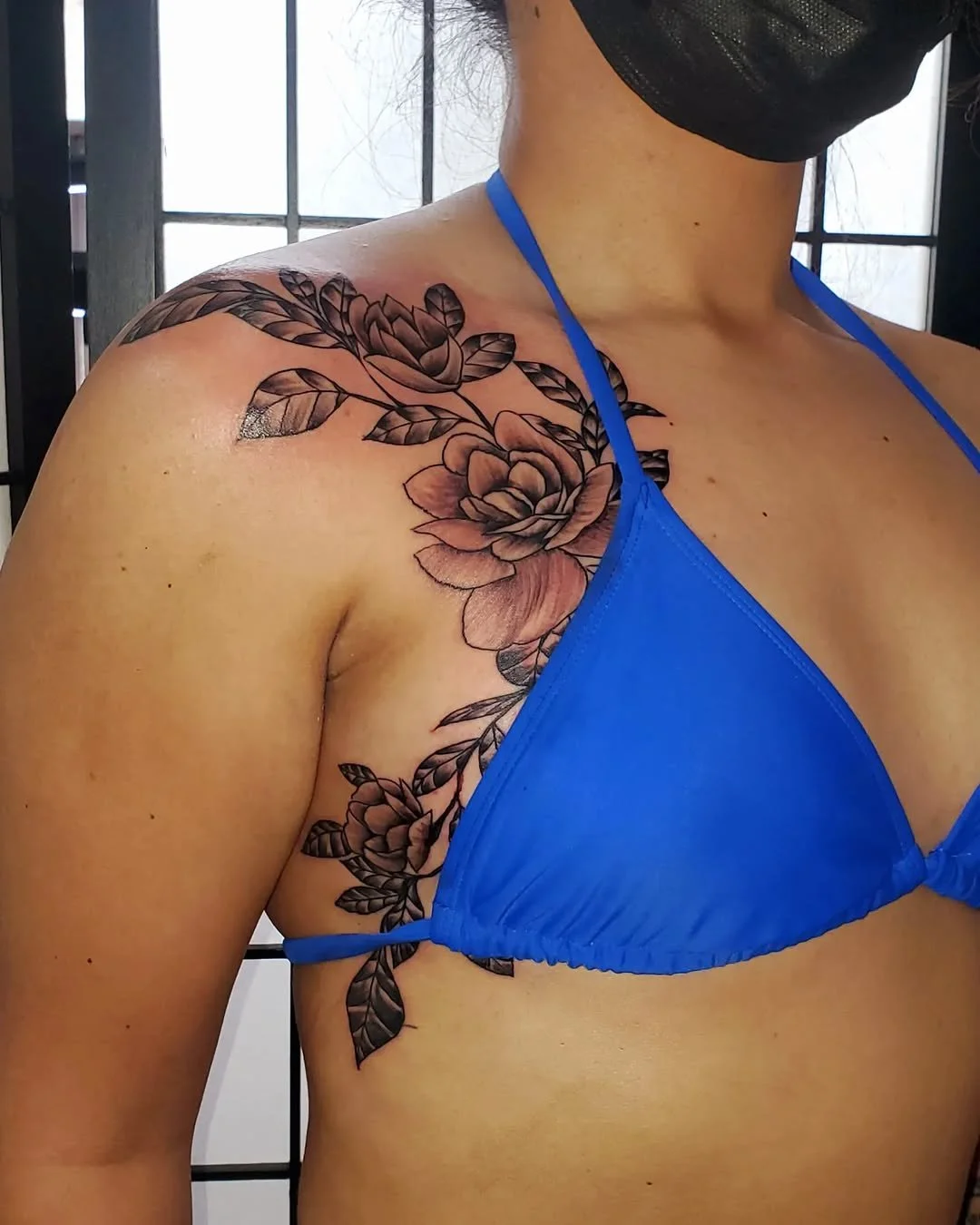 Close-up of a woman's shoulder tattoo of roses and leaves, wearing a blue bikini top and black face mask.
