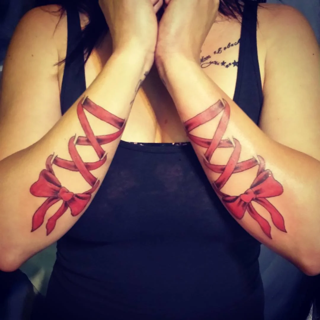 Close-up of a person's arms with matching tattoo designs of red ribbons forming a bow at the wrist, with the ribbons extending and wrapping around the forearms.