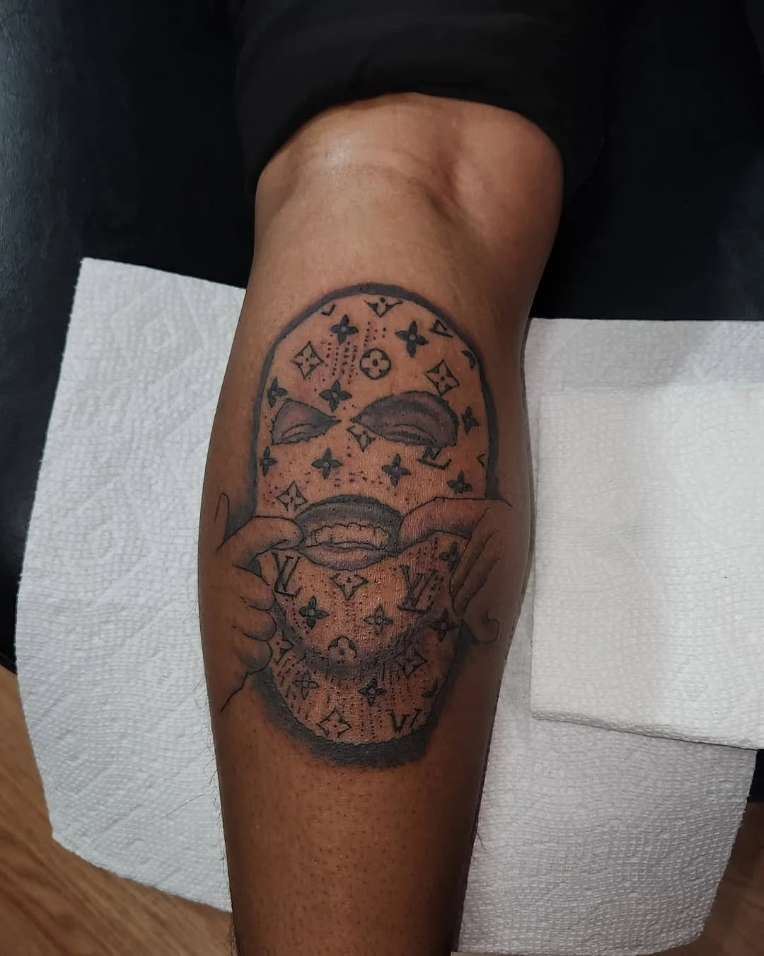 Tattoo of a face with lips pulling down a Louis Vuitton makeup compact, with the face's eyes closed and lips showing teeth, surrounded by Louis Vuitton logos.