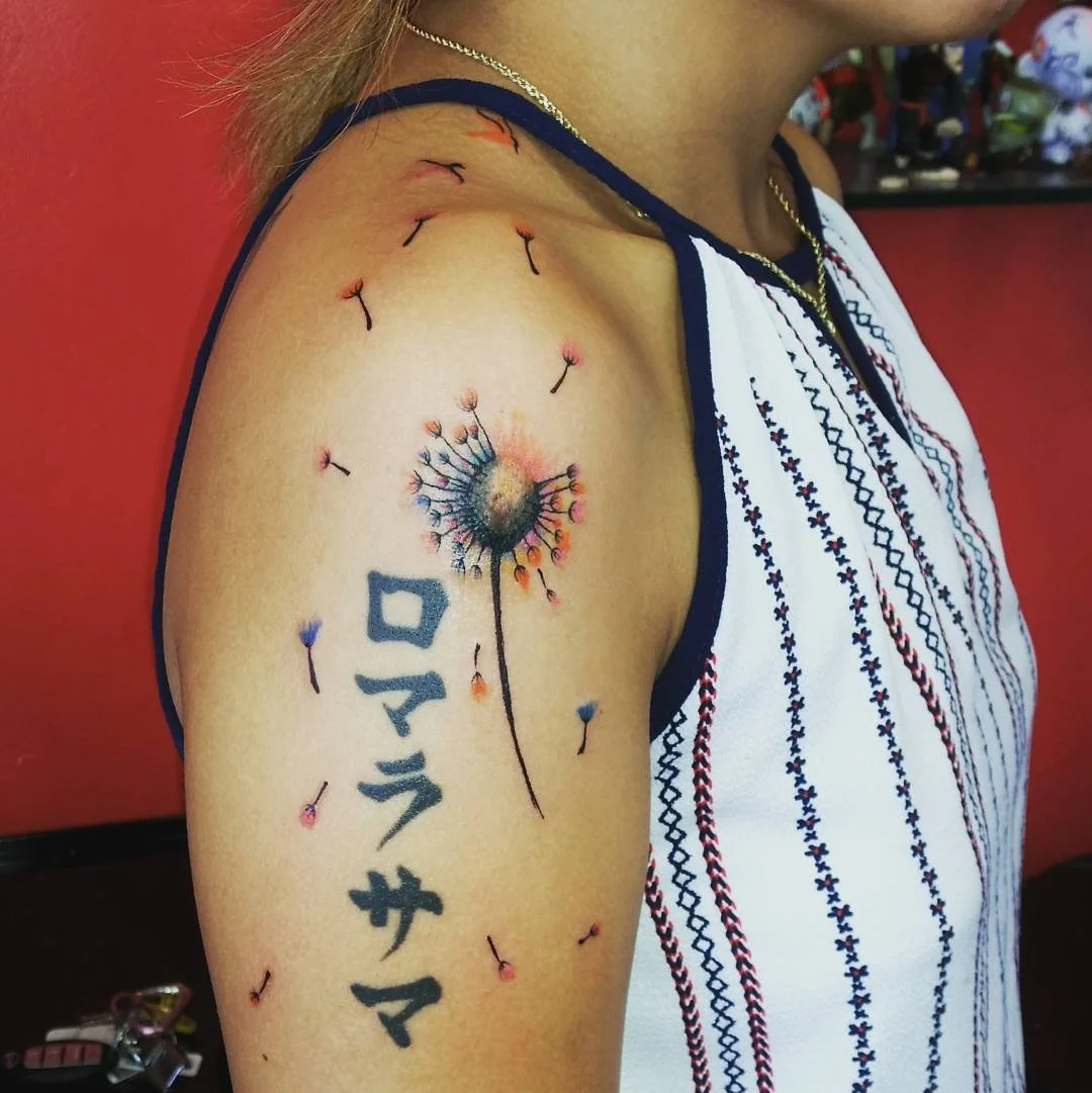A close-up of a person's shoulder with a dandelion tattoo and Japanese characters, wearing a sleeveless top with embroidery and a gold necklace.