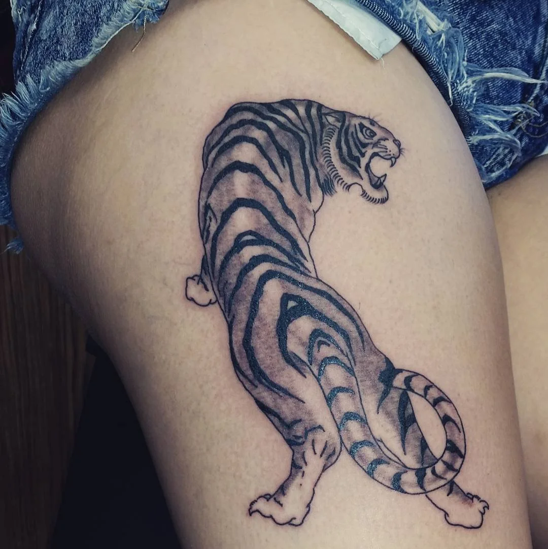 Tattoo of a roaring tiger with detailed black and gray stripes on a person's thigh.