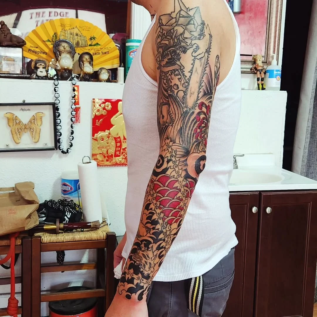 A person with a full sleeve tattoo on their left arm, featuring a warrior with a sword, waves, and colorful patterns, wearing a white tank top in an indoor setting with shelves, decorations, and a sink in the background.