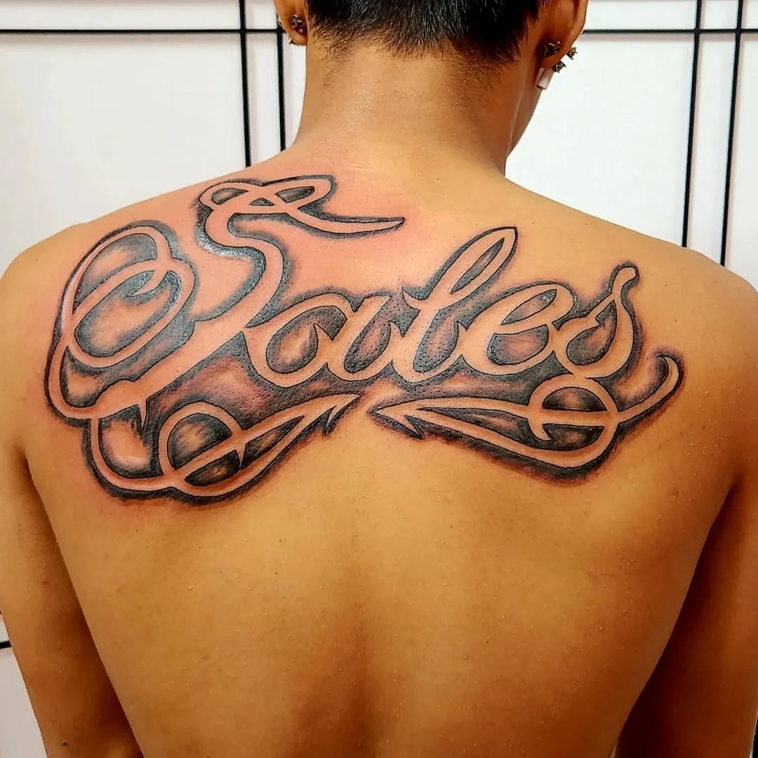 Tattoo of the word 'Loved' in cursive script on a person's upper back, with dark shading and bold lines.
