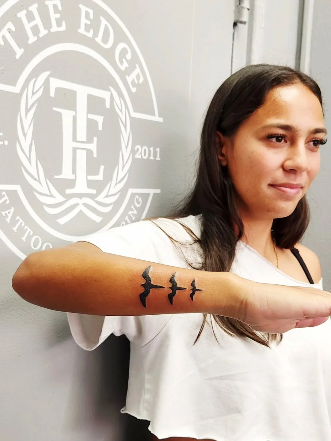 A woman shows a tattoo of three flying birds on her forearm, standing in front of a wall with a logo that reads "The Edge Tattoo Co." and the year "2011."