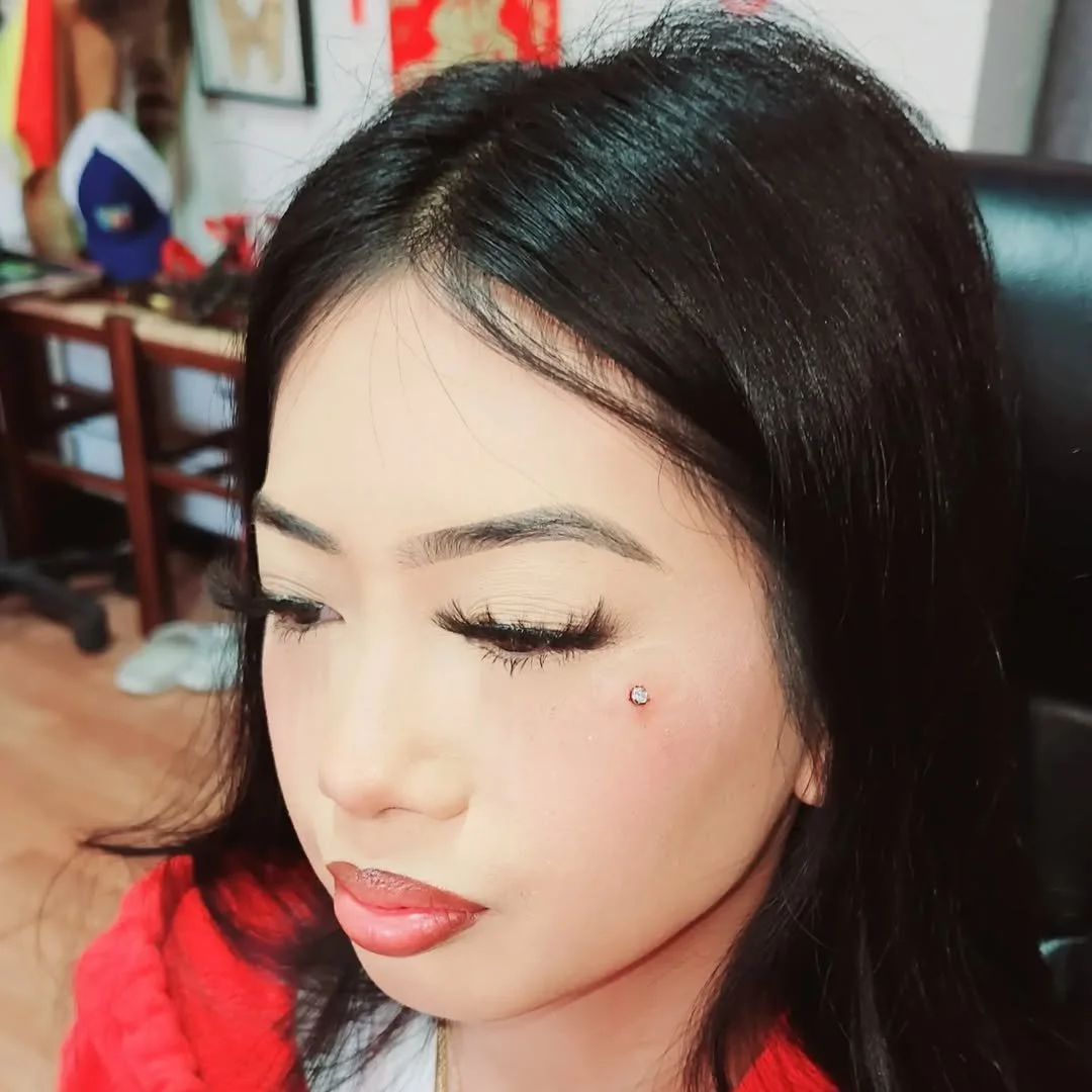 A woman with blonde eye makeup and long eyelashes, wearing a red top, has a small gemstone stud on her cheek.