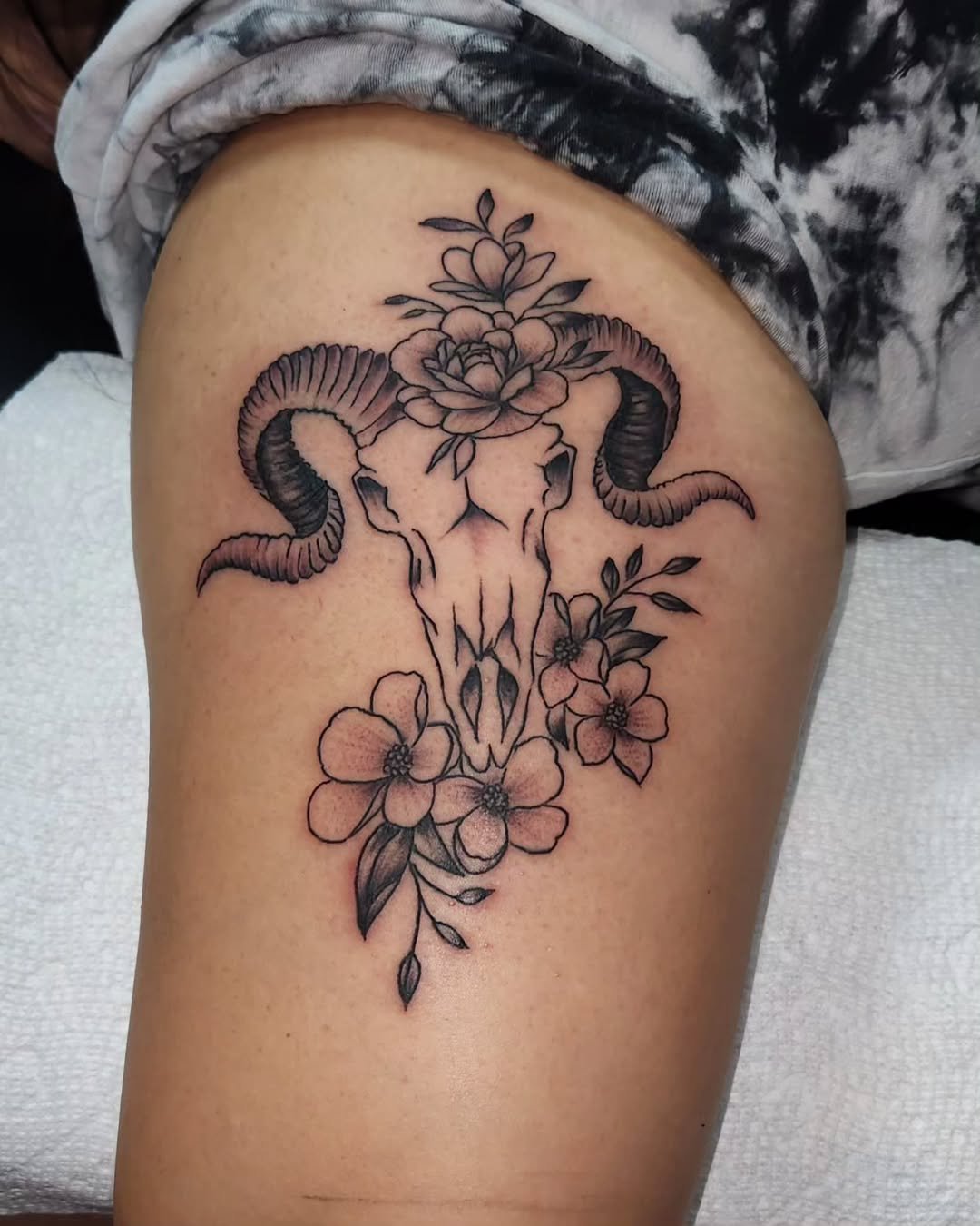 Tattoo of a ram skull with large curved horns, surrounded by flowers, on a person's thigh.