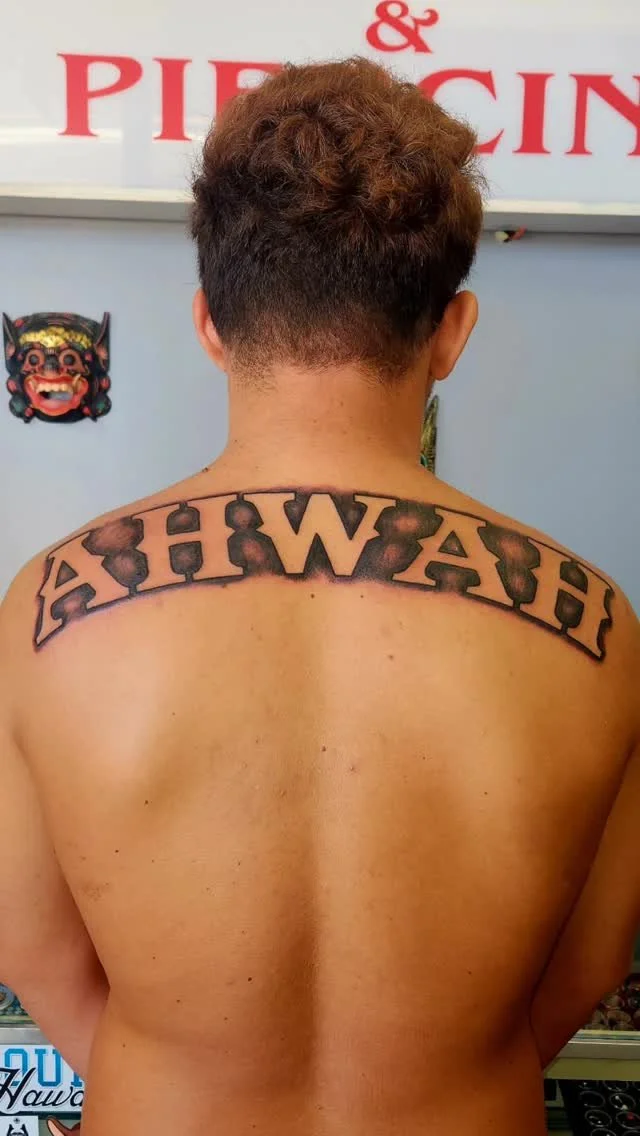 Back of a shirtless person with a tattoo on their upper back that reads 'A H I W A H!' in bold, black letters with a black outline.