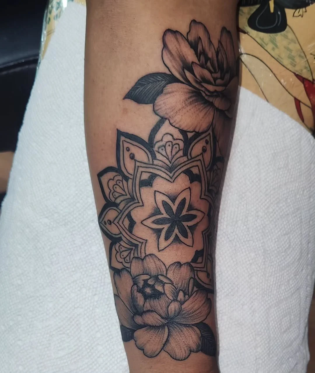 A tattoo featuring two large peony flowers with detailed petals and leaves, and a central mandala pattern with floral and geometric elements, all in black ink.