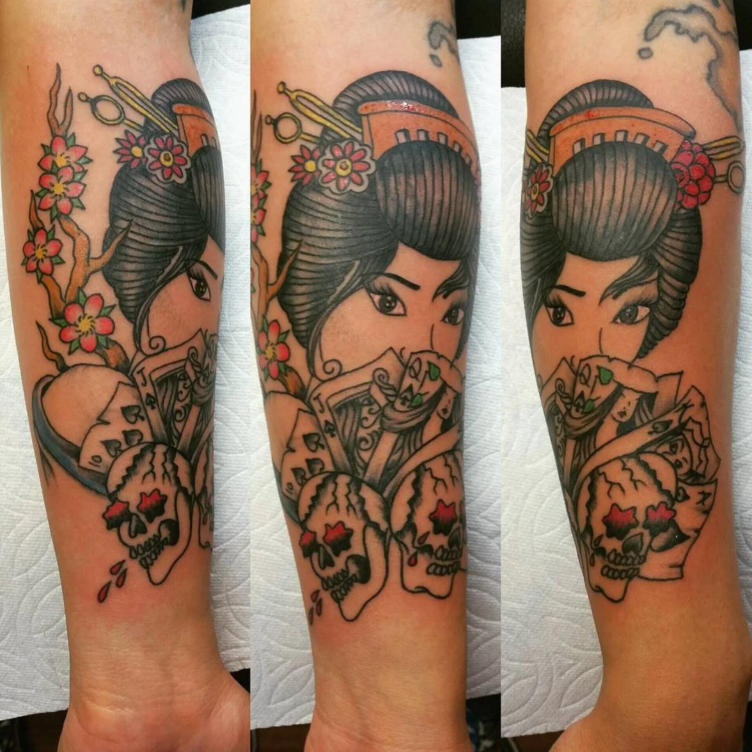 Tattoo of a woman with traditional hair and makeup, holding playing cards, with cherry blossoms and skulls at the bottom.