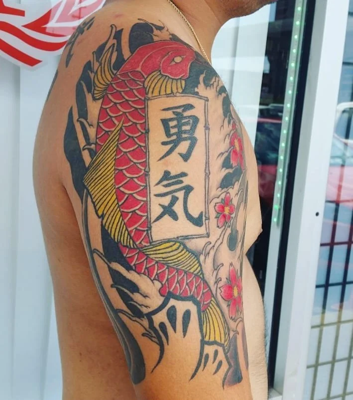 Tattoo of a red and yellow koi fish flowing around a rectangular sign with Japanese characters, surrounded by pink cherry blossoms.