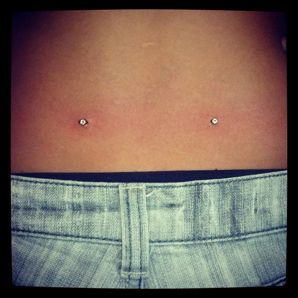 Close-up of a person's midsection with two belly button piercings and denim pants.